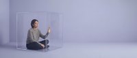 Young Asian woman in beige sweater sitting cross-legged inside transparent glass cube in minimalist purple studio space for meditation and mindfulness concepts.