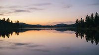 Serene lake at sunrise with forested mountains reflected in calm water, golden light illuminating sky and landscape for nature and travel backgrounds.