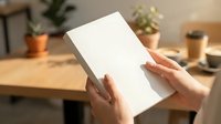 Female hands holding blank white notebook on wooden desk with potted plants and office supplies, suitable for branding, stationery mockups, and workspace design projects.