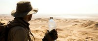 Man in cowboy hat holding water bottle in arid desert landscape, wearing backpack and dark clothing, suitable for travel, adventure, and outdoor survival themes.