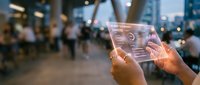 Hands holding transparent tablet displaying futuristic interface with holographic elements in modern urban environment with blurred city lights and people walking.
