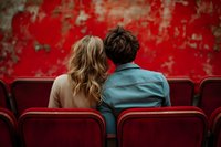 Young couple sitting close together in empty theater with red velvet seats and weathered crimson wall, viewed from behind, creating intimate romantic atmosphere.