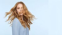 Young blonde Caucasian woman with flowing hair in motion wearing light blue sweater against white background for lifestyle and fashion content.
