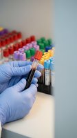 Medical laboratory technician in blue gloves holding blood sample test tube with colorful specimen collection tubes in background for clinical analysis.