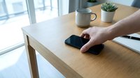 Man's hand holding smartphone on wooden desk with potted succulents, notebook and cup near bright window in modern office workspace.