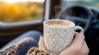 Steaming coffee in speckled ceramic mug held against car window during sunrise, cozy autumn morning atmosphere with warm drink and plaid blanket.