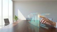 Business professional analyzing holographic data charts and graphs on wooden desk in modern office workspace with natural lighting and laptop computer.