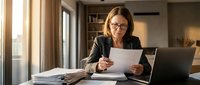 Professional mature businesswoman with glasses reviewing documents at modern office desk with laptop, focused on paperwork analysis and business planning.