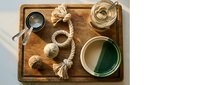 Overhead view of wooden tray with natural fiber rope, measuring cups, glass jars containing paint or liquid, and small bowls arranged on white surface for craft or DIY project.