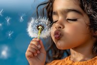 Young Hispanic girl with curly hair blowing dandelion seeds against blue sky background, wearing orange shirt, eyes closed in playful expression, childhood joy moment.