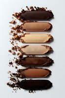 Crushed makeup powder swatches in various brown and nude shades arranged vertically on white background, showing texture and pigment intensity of cosmetic products.