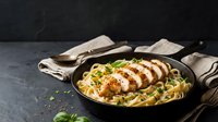 Grilled chicken breast sliced over creamy pasta with herbs in cast iron skillet on dark slate background for food photography and culinary content.
