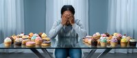 Young Asian man in gray hoodie covering face with hands sitting at table full of colorful cupcakes and donuts showing diet temptation and sugar addiction struggle.