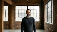 Confident middle-aged Caucasian businessman in dark sweater standing in modern industrial loft office with brick walls and large windows, professional portrait.