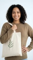 Young woman with curly black hair and medium skin tone smiling while holding organic cotton tote bag with leaf design, wearing tan ribbed sweater for eco-friendly lifestyle and sustainable shopping concepts.