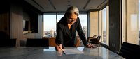Senior businesswoman with gray hair and glasses leaning over marble desk in modern office with city skyline view, wearing black blazer, professional executive portrait.