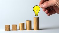 Hand placing yellow lightbulb on ascending wooden blocks showing growth, innovation and business progress concept on light background.