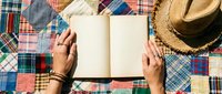 Hands holding open blank notebook on colorful patchwork quilt background with straw hat. Perfect for journaling, writing, planning and creative projects.