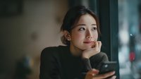 Young Asian businesswoman smiling while using smartphone near office window, professional female entrepreneur checking mobile phone during work break.