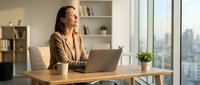 Professional businesswoman in beige blazer working on laptop at modern office desk with city view, contemplating business strategy and planning.