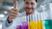 Young male scientist in lab coat and glasses showing thumbs up while working with colorful liquid samples in test tubes at modern laboratory setting.