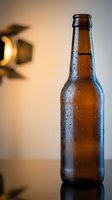Cold brown glass beer bottle with water droplets on dark surface with warm lighting for beverage advertising and restaurant marketing materials.