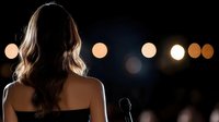 Silhouette of woman with long wavy hair from behind facing stage with microphone and blurred bokeh lights in dark background.