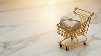 Miniature golden shopping cart filled with white gift box on marble surface, representing retail commerce and consumer spending.