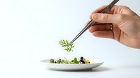Chef's hand using tweezers to plate a gourmet microgreen garnish on an elegant white dish, fine dining presentation on white background.
