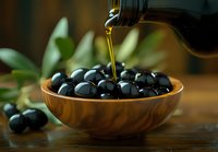 Fresh olive oil pouring from bottle onto black olives in wooden bowl, with olive leaves and scattered olives on rustic table.