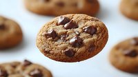 Freshly baked chocolate chip cookie floating in air against white background, showcasing golden brown texture and melty chocolate chunks.