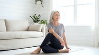 Mature woman with gray hair, Caucasian ethnicity, sitting on yoga mat in bright living room practicing mindfulness and meditation for wellness and mental health.