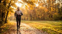 Senior man jogging through autumn park with golden sunlight filtering through colorful fall foliage creating warm atmospheric morning exercise scene.