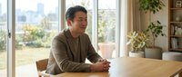 Professional middle-aged Asian businessman in casual brown sweater sitting at wooden table in bright modern office with large windows and city view.