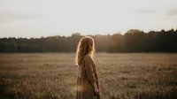 Young woman with long wavy hair standing in golden field during sunset, peaceful countryside moment with warm natural lighting and serene atmosphere.