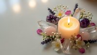 Burning candle surrounded by delicate flowers, rose petals and crystals creating peaceful spa atmosphere for relaxation and wellness rituals.