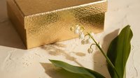 Luxury golden gift box with lily of the valley flowers on textured cream background for premium packaging and elegant present concepts.
