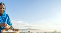 Senior woman wearing blue headscarf holding basket of fresh fish at dawn, mountains visible in misty background. Suitable for agriculture, fishing industry, and cultural lifestyle content.