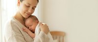 Young Asian mother in beige sweater tenderly holding sleeping newborn baby in bright nursery room. Peaceful maternal bonding moment for parenting content.