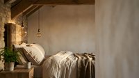 Rustic bedroom with exposed wooden beams, textured plaster walls, and warm pendant lighting above an unmade bed with neutral linens and throw blanket.