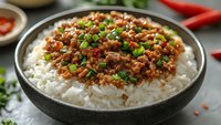 Spicy minced meat with green onions served over steamed white rice in black ceramic bowl, garnished with chili peppers and fresh herbs for Asian cuisine.