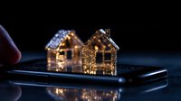 Glowing miniature houses on smartphone screen in dark room create magical holiday atmosphere with warm golden lights and bokeh effect.