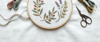 Embroidery hoop with botanical floral design featuring leaves and branches, surrounded by colorful threads and scissors on white fabric background.