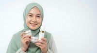 Young Muslim woman in green hijab holding skincare cream jar and cosmetic bottle, smiling at camera on white background for beauty and wellness marketing.