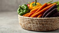 Fresh organic vegetables in woven basket including vibrant carrots, purple carrots, green and yellow bell peppers on neutral background.