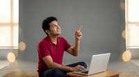 Young man with dark hair and medium skin tone, wearing a burgundy polo shirt, sitting at wooden desk with laptop, pointing upward with one finger while looking to the side in bright office space with windows.