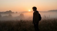 Young man with backpack standing in misty field at sunrise, silhouette against golden sky, peaceful morning landscape with fog rolling over countryside.