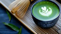 Hot green matcha latte with white foam art heart design in black ceramic cup, bamboo whisk and fresh tea leaves on wooden mat, close up view.