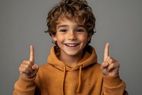 Young boy with curly brown hair wearing orange hoodie showing peace signs with both hands while smiling cheerfully against gray studio background.