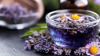 Purple lavender tea in glass bowl with fresh flowers and chamomile blooms on dark stone background, aromatic herbal drink for relaxation and wellness.